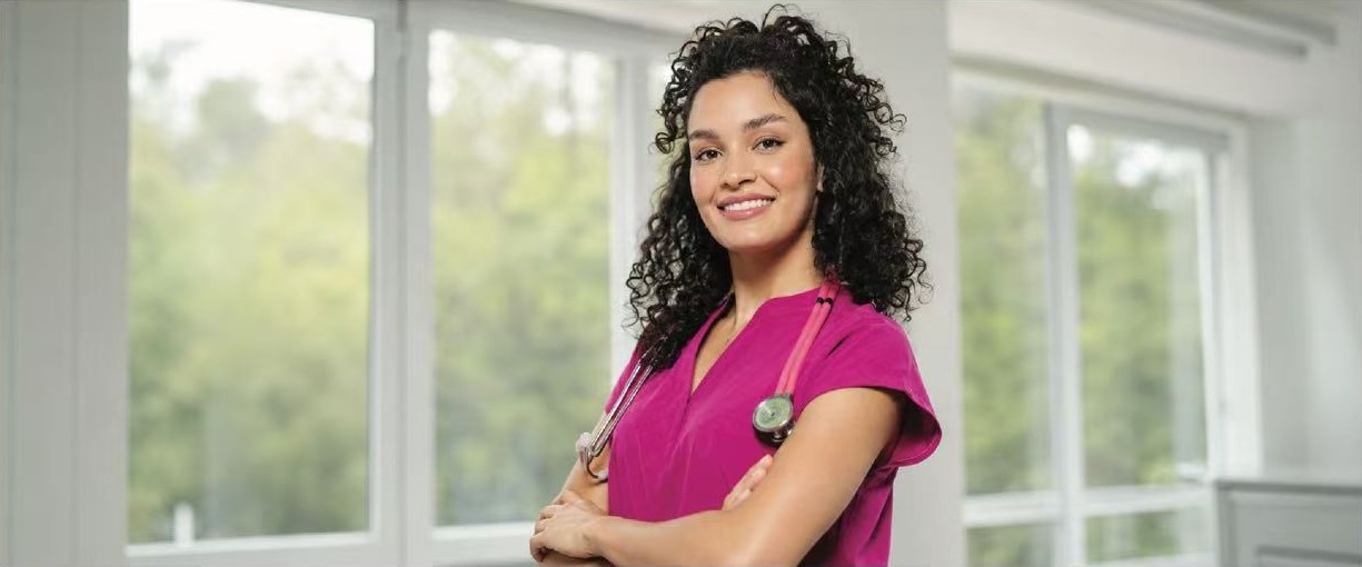 Healthcare professional in pink scrubs standing near bright windows